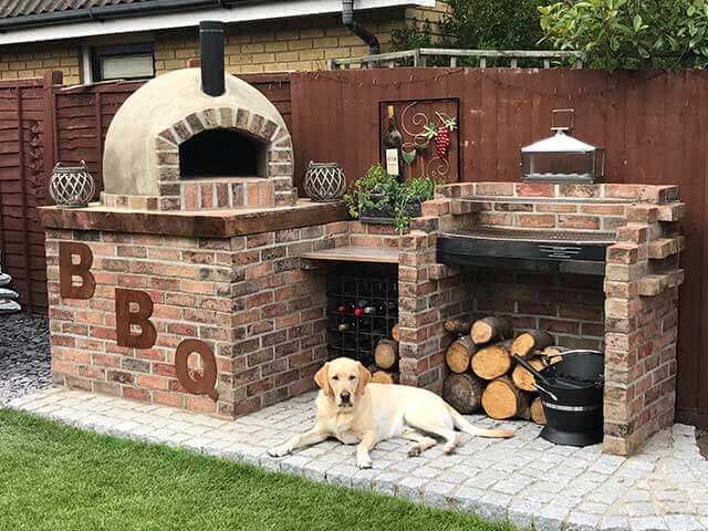 A cost-effective yet aesthetically decorated pizza oven and kitchen set up made from bricks in a back garden with a labrador sitting before it all in the sun.